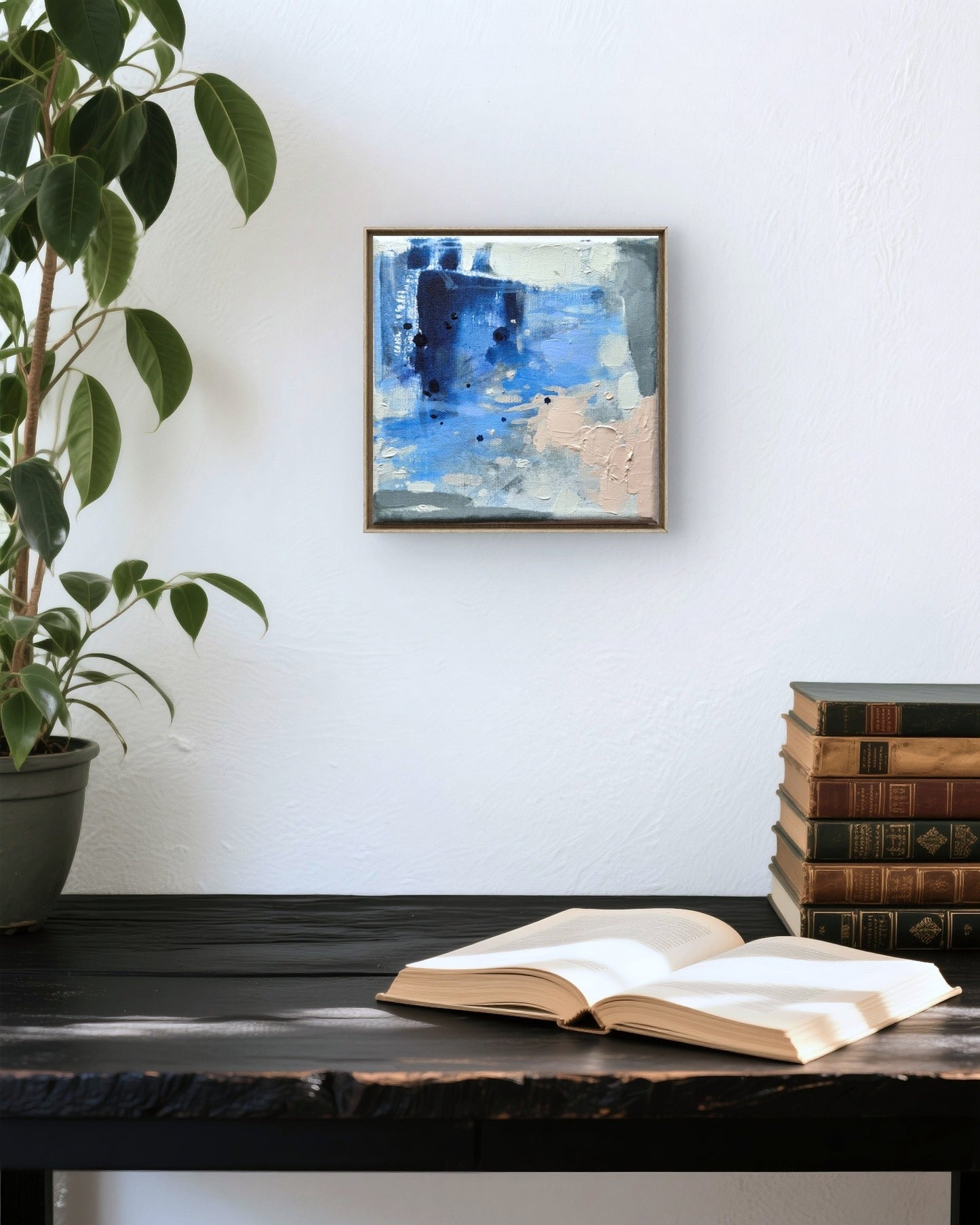 A small framed abstract painting with blue, white, and beige tones hangs on a white wall above a dark wooden table. An open book rests on the table, with a stack of vintage books and a potted plant visible in the scene.