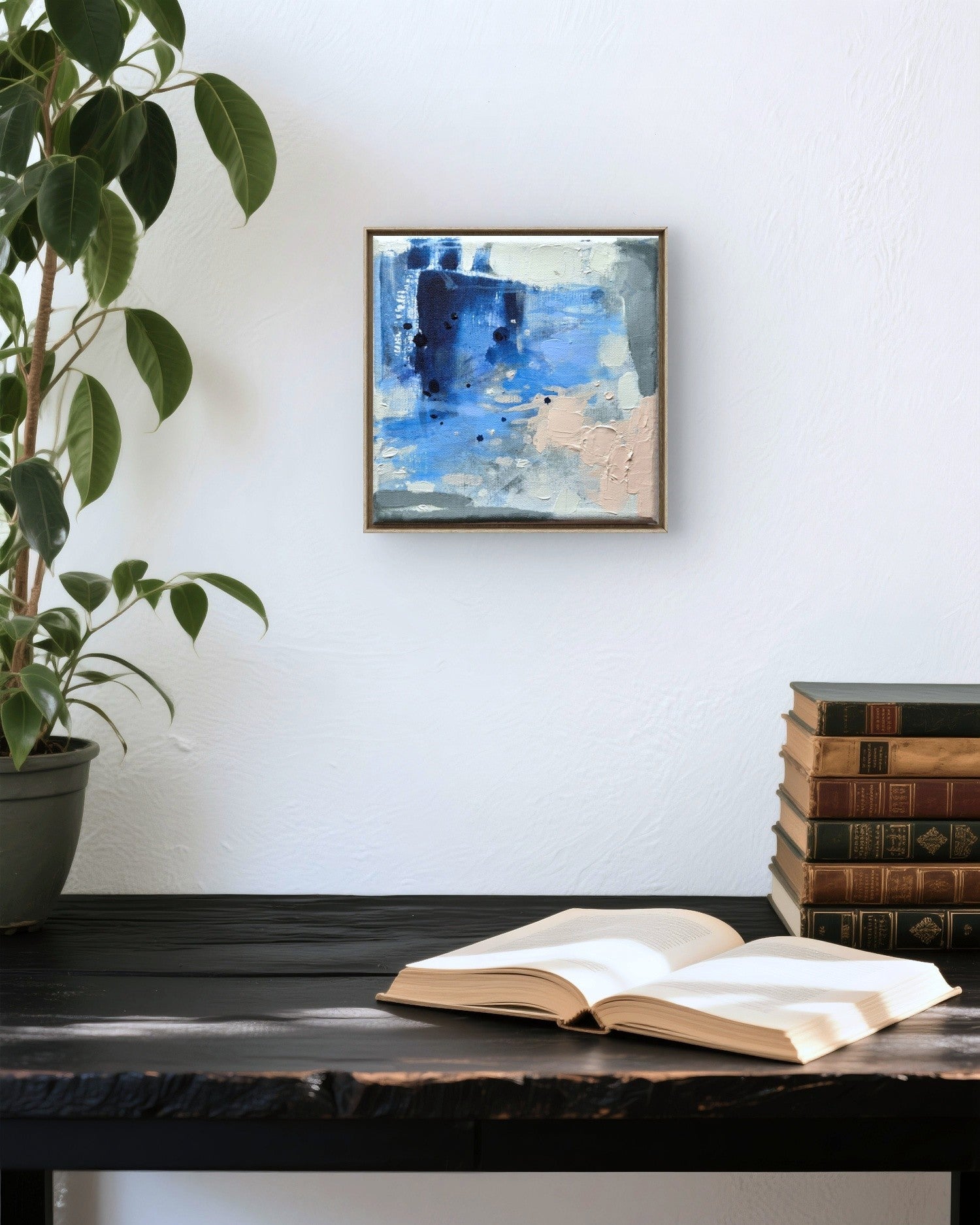A small framed abstract painting with blue, white, and beige tones hangs on a white wall above a dark wooden table. An open book rests on the table, with a stack of vintage books and a potted plant visible in the scene.