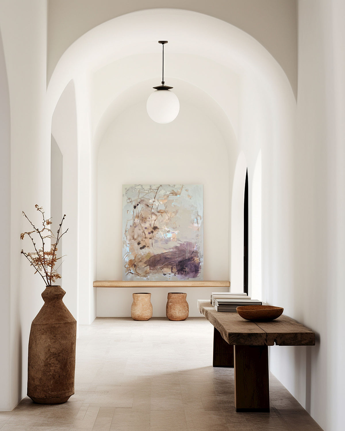 A modern architectural hallway with white arches and walls, featuring a rustic wooden bench, a large brown ceramic vase with dried branches, a small abstract painting, and a spherical white pendant light.