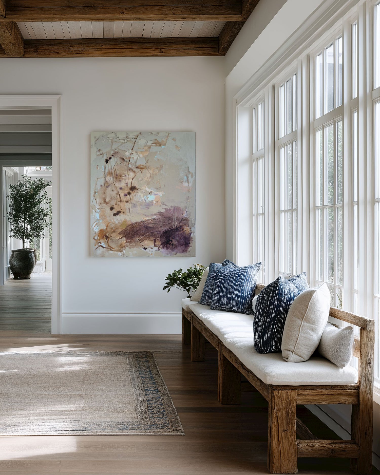 An abstract canvas painting with soft beige, brown, and purple brushstrokes hangs on a white wall in a bright, modern room. Below the painting is a wooden bench with a white cushion and blue pillows, next to tall white-framed windows.