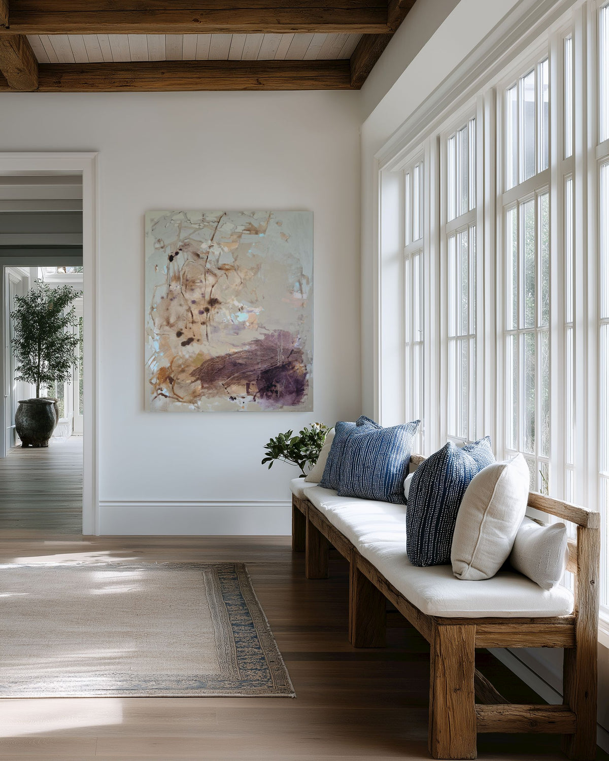 An abstract canvas painting with soft beige, brown, and purple brushstrokes hangs on a white wall in a bright, modern room. Below the painting is a wooden bench with a white cushion and blue pillows, next to tall white-framed windows.