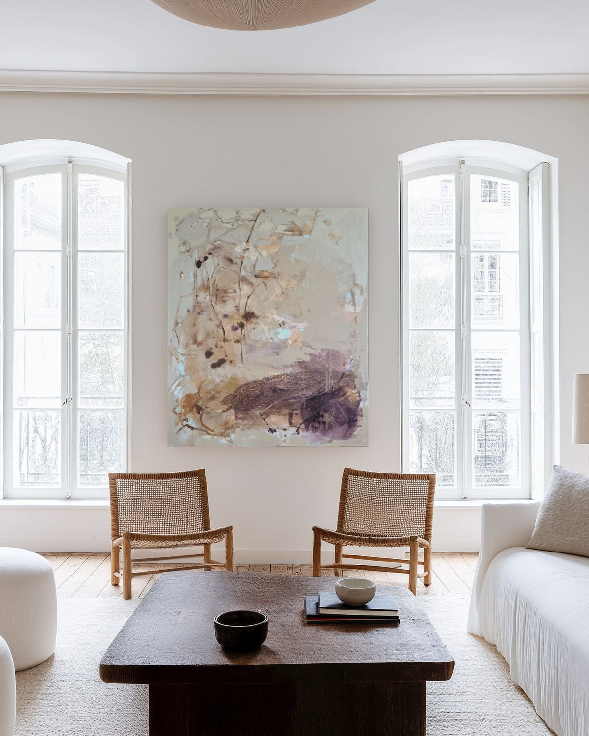 A bright, modern living room with white walls, two tall arched windows, a white sofa, two wooden chairs, a dark wood coffee table, and two large abstract wall art pieces hanging on the walls.