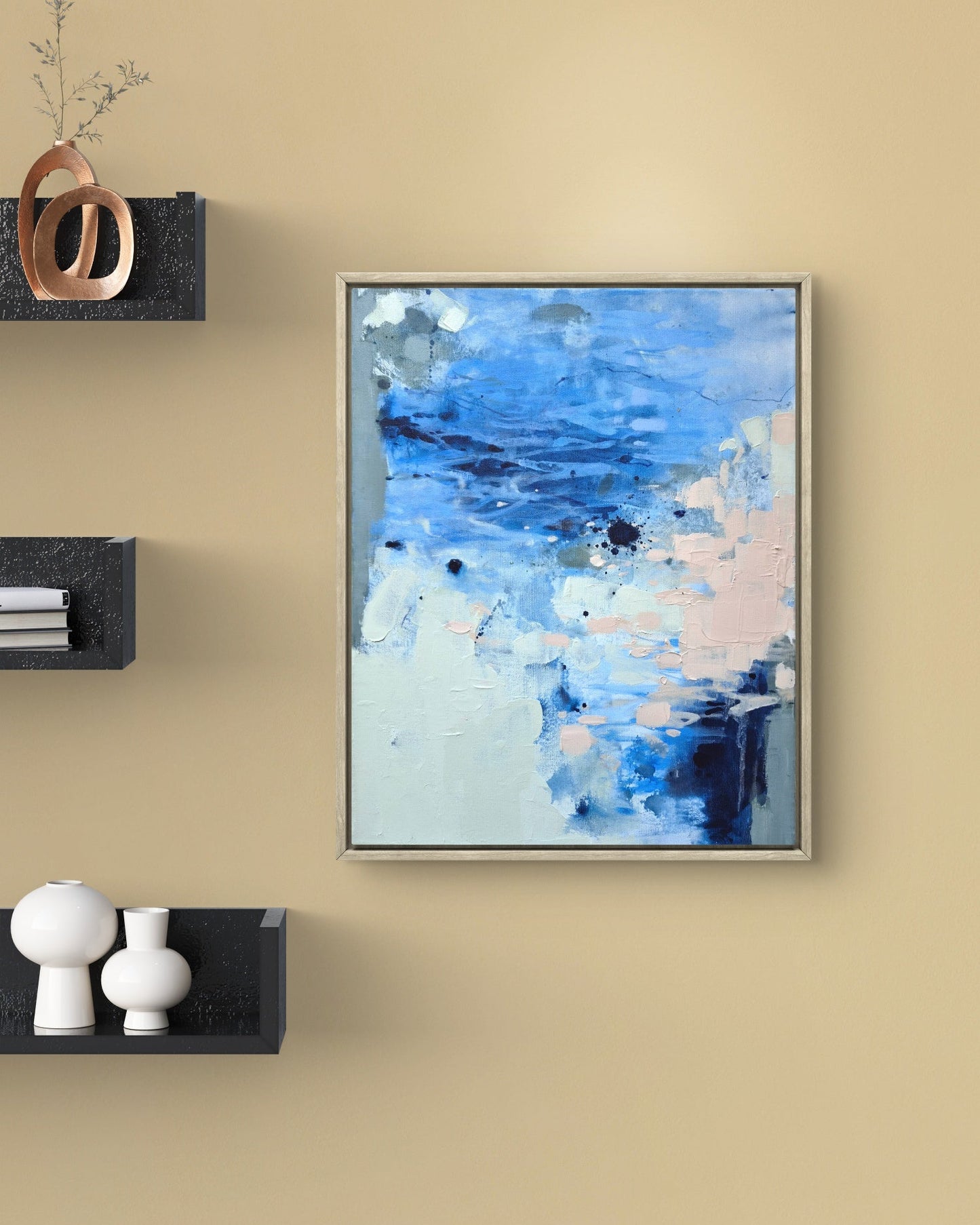 An abstract framed painting with blue, white, and soft pink brushstrokes, displayed on a beige wall with floating black shelves holding decorative objects.