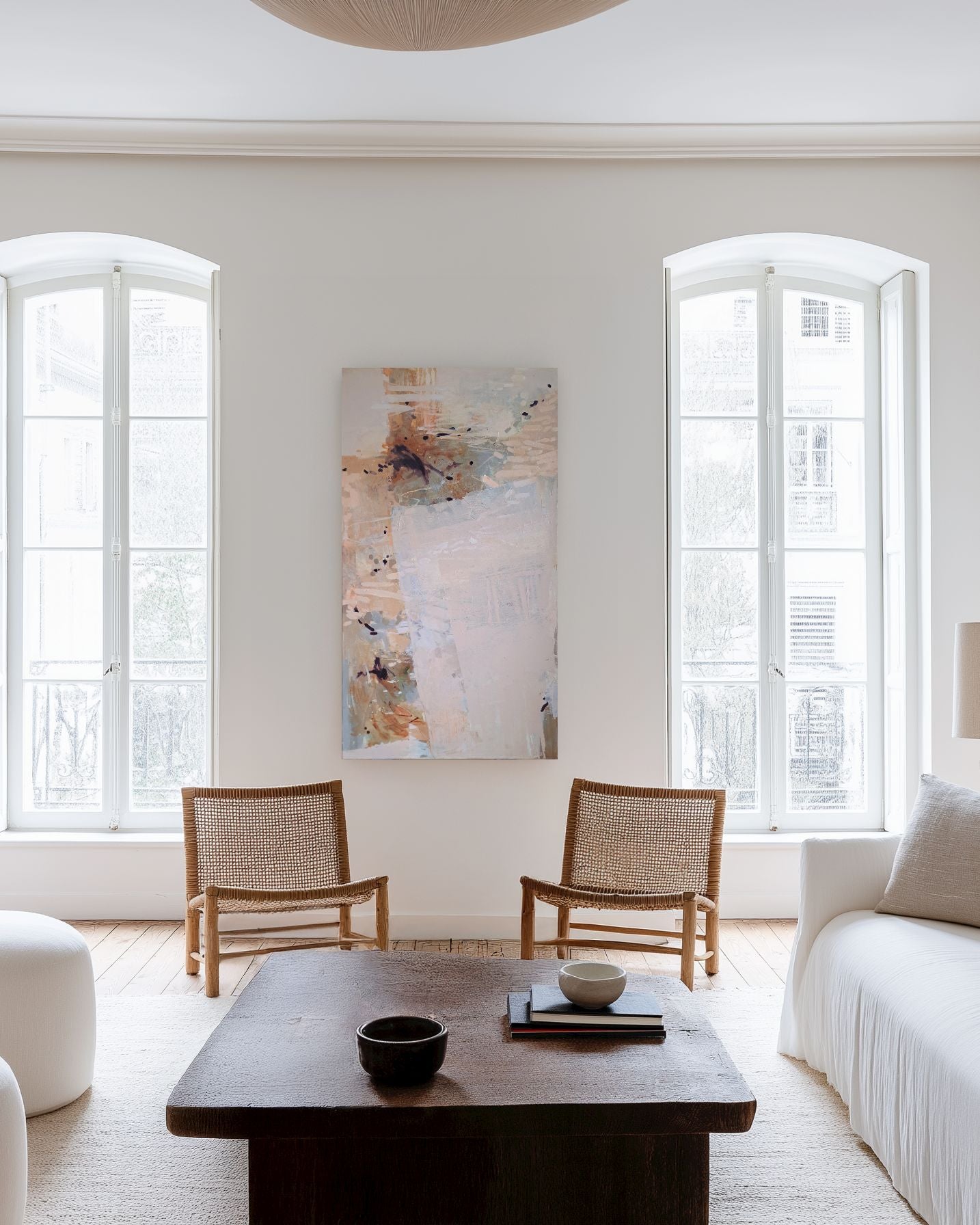 An abstract canvas painting with soft beige, white, and brown tones hangs on a white wall in a bright, modern living room with two wooden armchairs, a dark wood coffee table, and white upholstered seating.