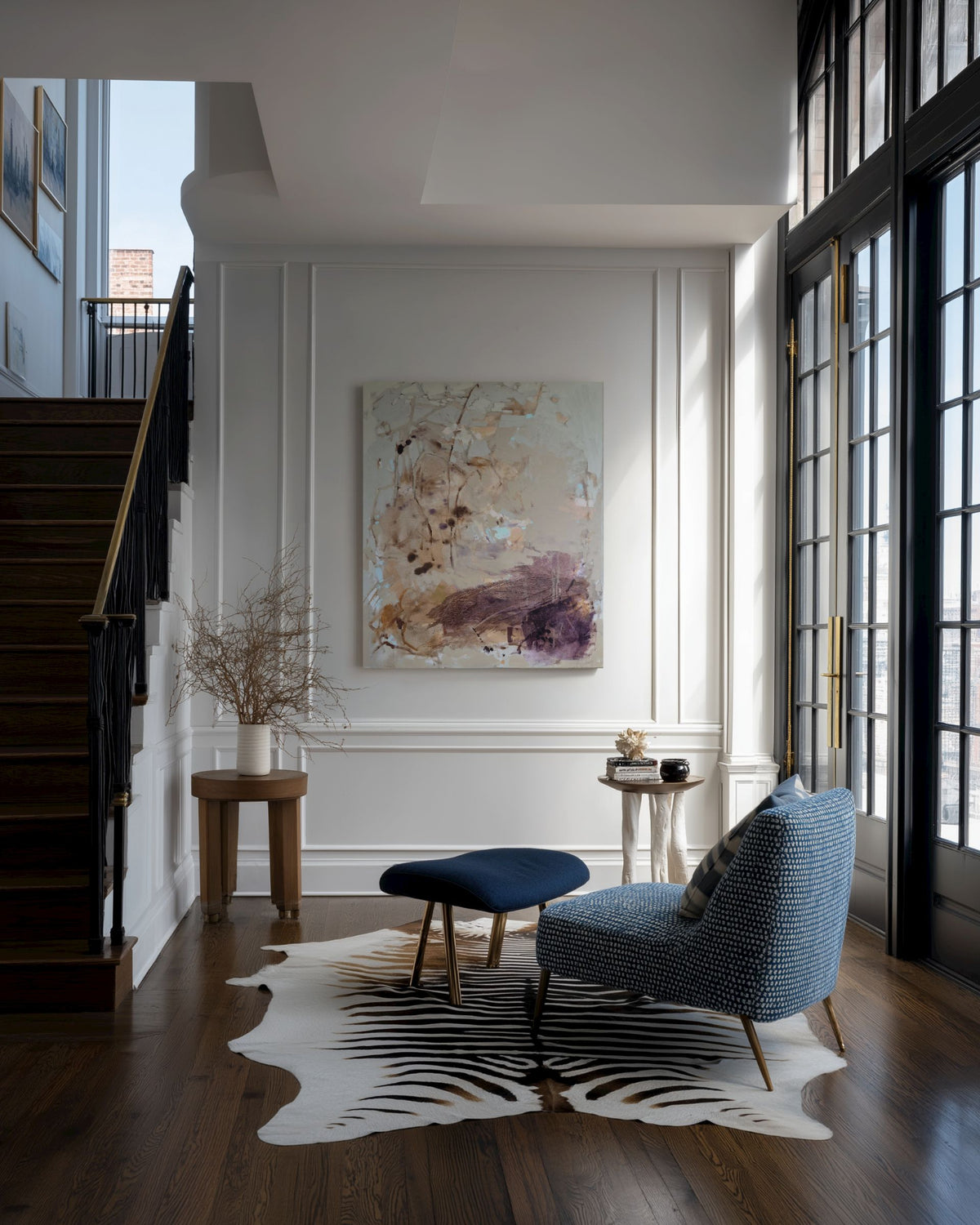 A styled interior entryway with a blue upholstered armchair, a matching blue ottoman, a black-and-white cowhide rug, a wooden staircase, and a large abstract painting on a white wall.