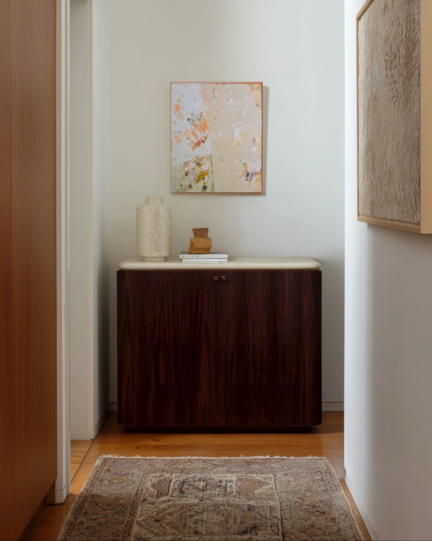 Narrow hallway with a wooden cabinet, decorative items, and framed artwork on white walls.