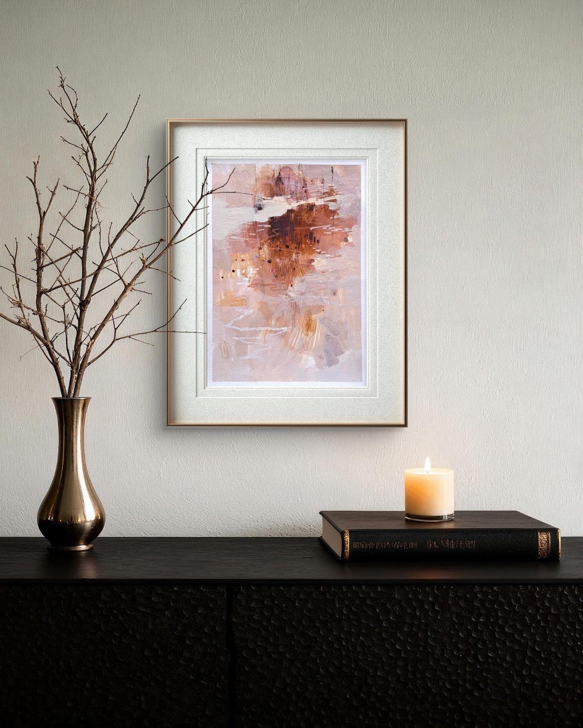 Framed abstract art piece on a wall above a dark surface with a vase, candle, and book.
